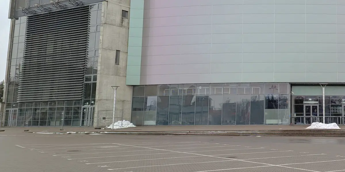 Išlenkta pilkšvai rausvų plokščių arenos fasadas su stikliniu įėjimų pirmu aukštu ir betoniniu bokštu kairėje; priekyje – didelė grįsta automobilių aikštelė su baltu ženklinimu.
