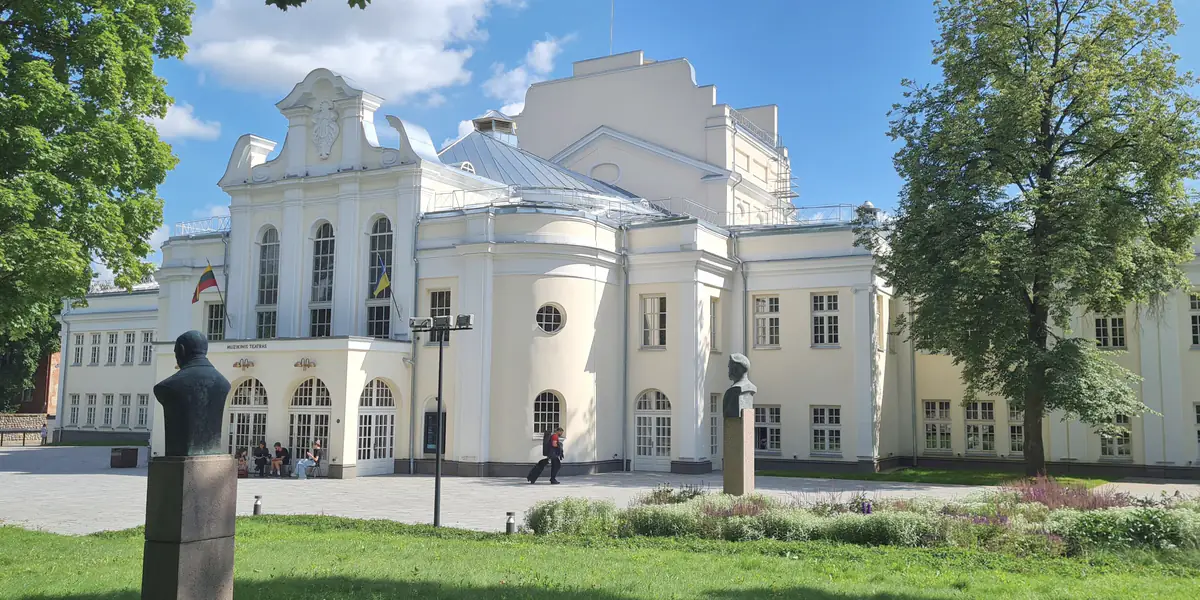Šviesus Kauno muzikinio teatro pastatas su arkiniais langais ir užrašu „Muzikinis teatras“ virš įėjimo; priekyje – teatro sodo veja ir dvi biustų skulptūros.