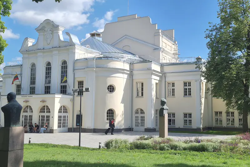 Šviesus Kauno muzikinio teatro pastatas su arkiniais langais ir užrašu „Muzikinis teatras“ virš įėjimo; priekyje – teatro sodo veja ir dvi biustų skulptūros.
