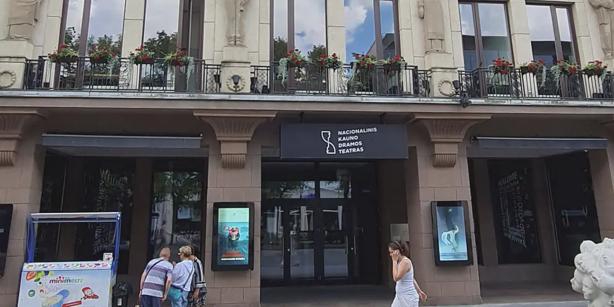 Nacionalinio Kauno dramos teatro įėjimas Laisvės alėjoje: juoda iškaba su teatro pavadinimu, ekranai su spektaklių afišomis, virš jų – balkonas su raudonomis pelargonijomis.
