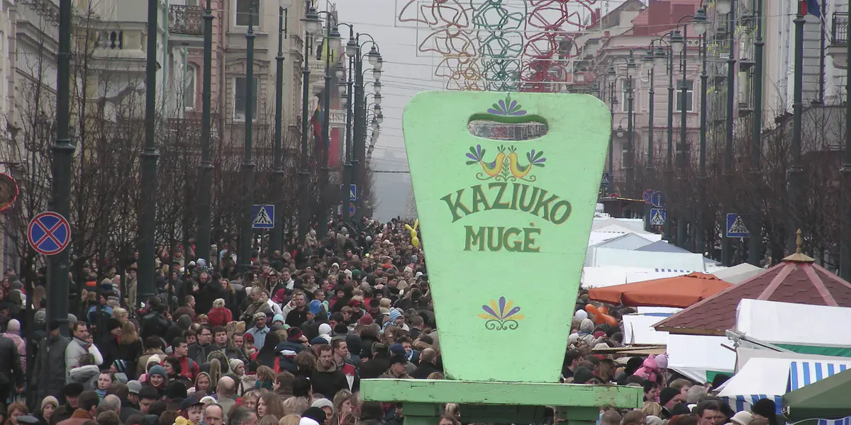 Kaziuko mugė Gedimino prospekte Vilniuje: pilna žmonių gatvė, viduryje – didelė žalia medinė verpstė su užrašu „Kaziuko mugė“, dešinėje – prekybos palapinės.