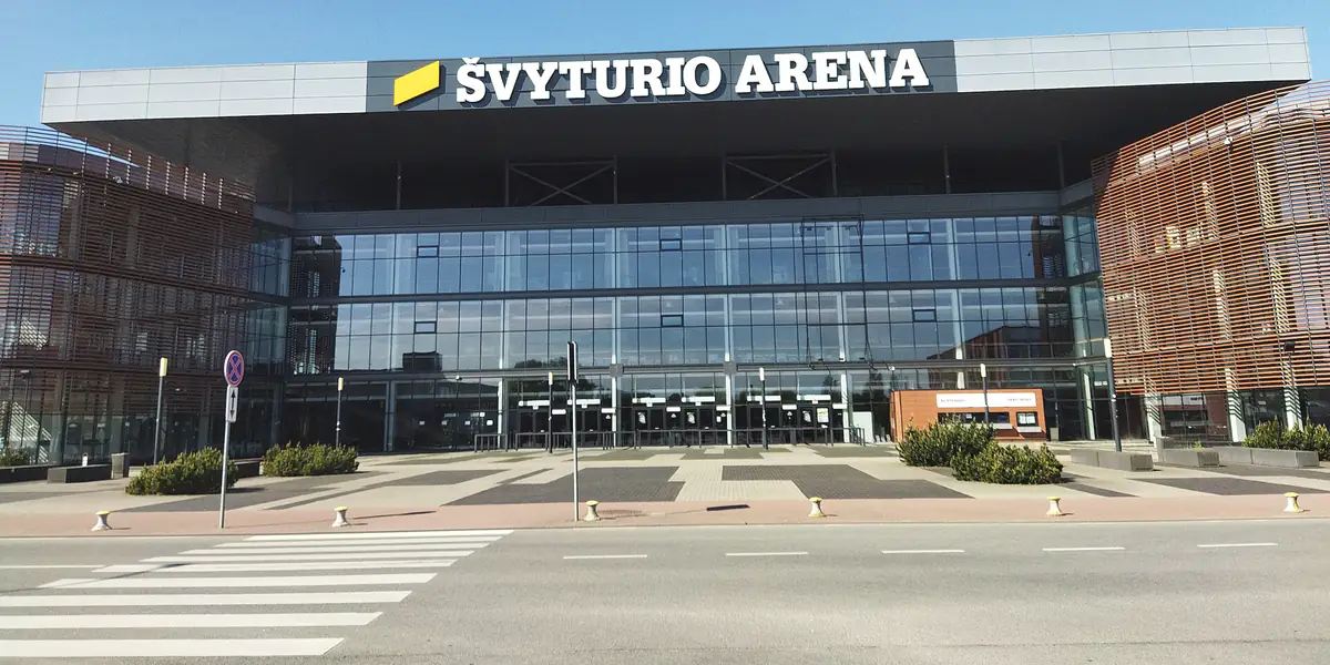 Stiklinis Švyturio arenos fasadas su užrašu „Švyturio arena“ ir geltonu ženklu virš įėjimo; priekyje – aikštė su krūmų grupėmis ir gatvės pėsčiųjų perėja.