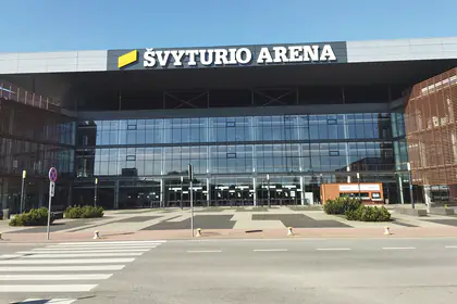 Stiklinis Švyturio arenos fasadas su užrašu „Švyturio arena“ ir geltonu ženklu virš įėjimo; priekyje – aikštė su krūmų grupėmis ir gatvės pėsčiųjų perėja.